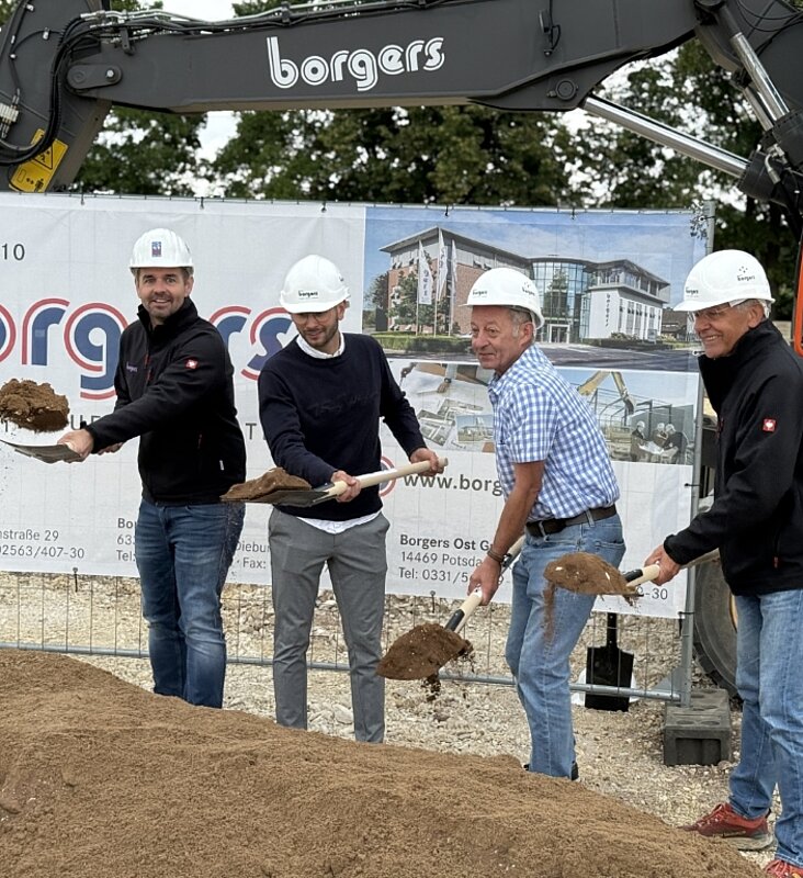 Vier Personen mit weißen Bauhelmen und Spaten in der Hand während eines Spatenstichs