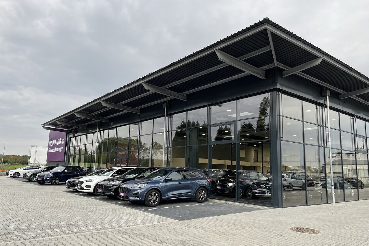 Modernes AGVV Autohaus mit Glasfront und gepflastertem Vorplatz, auf dem mehrere Neuwagen stehen