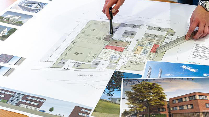 Blick von oben auf einen Tisch mit Bauplänen, Grundrissen und Skizzen, auf denen mit einem Stift gezeigt wird