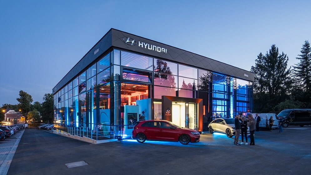 Abendliche Außenansicht eines beleuchteten Autohauses mit Glasfassade und einem roten Sportwagen