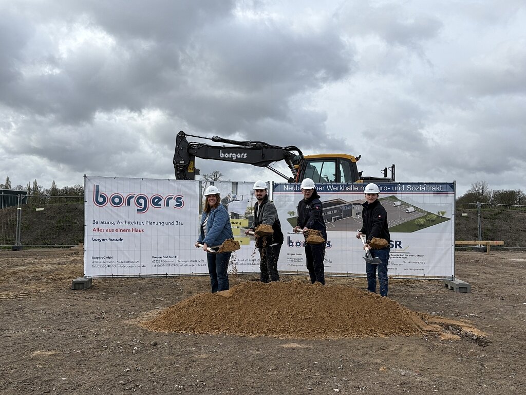 Vier Personen mit Spaten in der Hand beim Spatenstich, Bagger im Hintergrund zu sehen