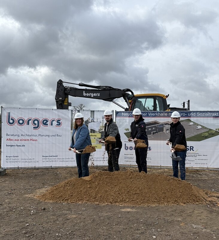Vier Personen mit Spaten in der Hand beim Spatenstich, Bagger im Hintergrund zu sehen