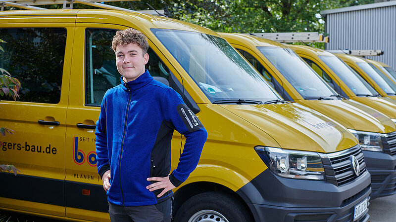 Maurer-Azubi der Borgers GmbH in blauer Jacke, der vor einer Reihe gelber Transporter steht