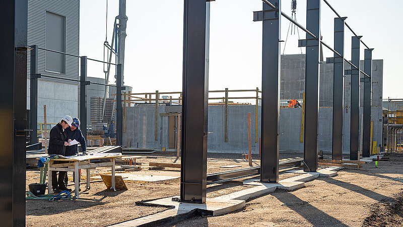 Innenansicht einer großen Baustelle im Rohbau, bei der Stahlträger für eine Gewerbehalle aufgestellt werden