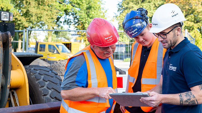 Drei Bauarbeiter in Bauhelmen und Warnwesten betrachten Baupläne auf einem Tablet