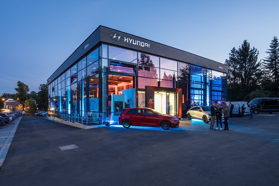 Abendliche Außenansicht eines beleuchteten Autohauses mit Glasfassade und einem roten Sportwagen