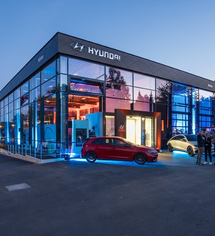 Abendliche Außenansicht eines beleuchteten Autohauses mit Glasfassade und einem roten Sportwagen