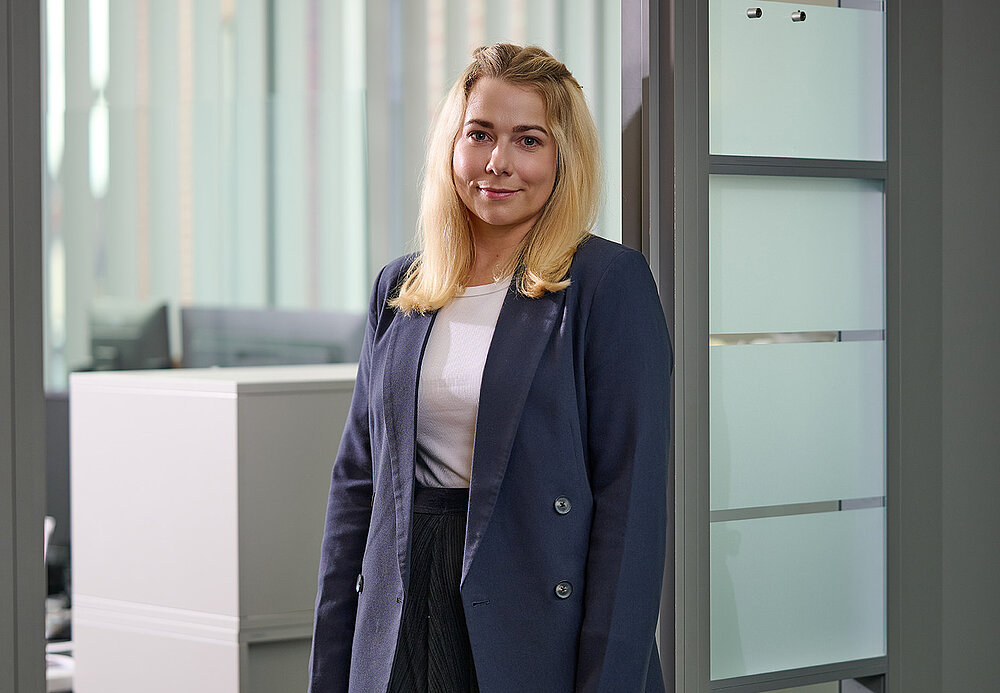 Portrait einer lächelnden jungen Frau in dunklem Blazer vor gläserner Bürotür