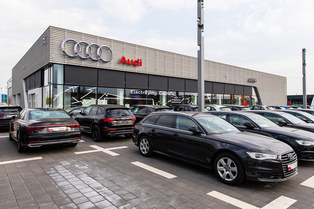 Außenansicht eines modernen Autohauses mit Verkaufsbereich und vielen E-Autos auf dem Gelände