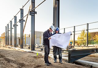 Zwei gewerbliche Mitarbeiter der Borgers GmbH auf der Baustelle. Sie schauen gemeinsam auf einen Plan. Im Hintergrund ist Stahlbau zu sehen.