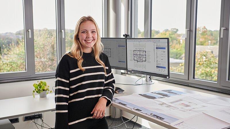 Junge Frau steht am Schreibtisch. Es sind Architekturpläne zu sehen