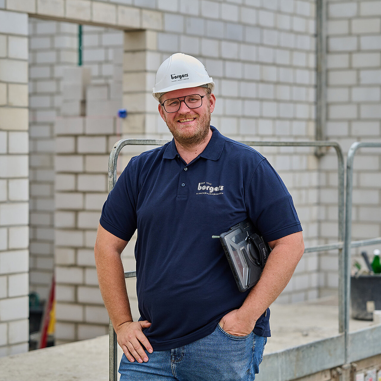 Mitarbeiter der Borgers GmbH in Bauhelm und Polo-Shirt, der auf einer Baustelle vor einem Ziegelrohbau posiert