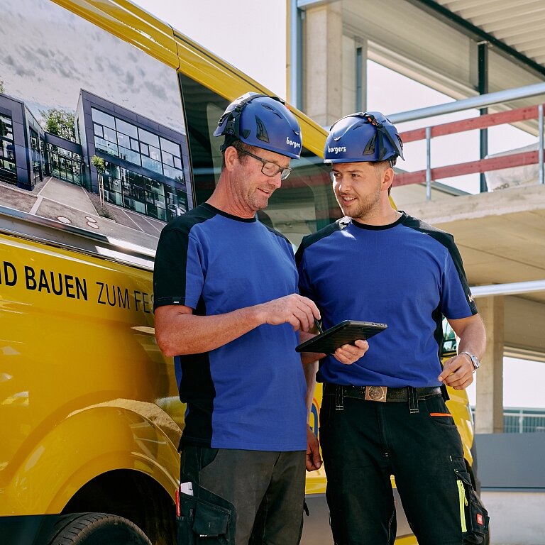 zwei gewerbliche Kollegen im Gespräch auf der Baustelle vor einem gelben Borgers Bulli mit Tablet in der Hand