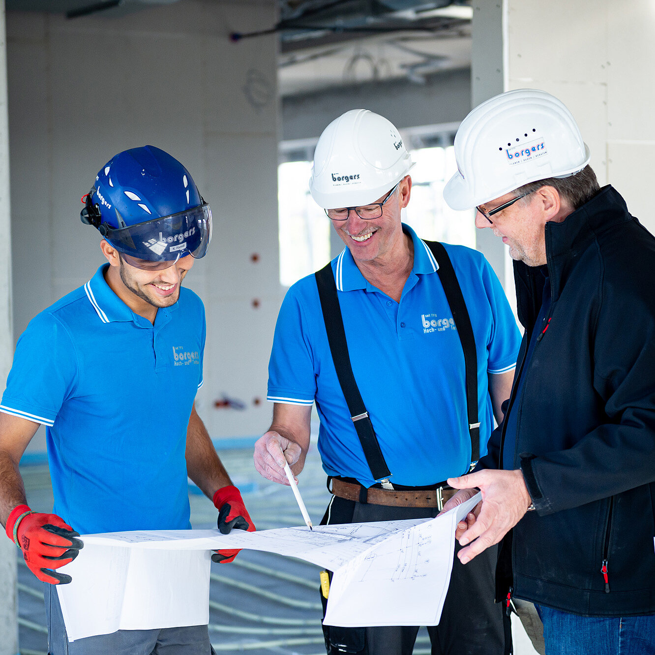 Drei Bauarbeiter in blauen T-Shirts und Helmen besprechen Baupläne auf einer Baustelle