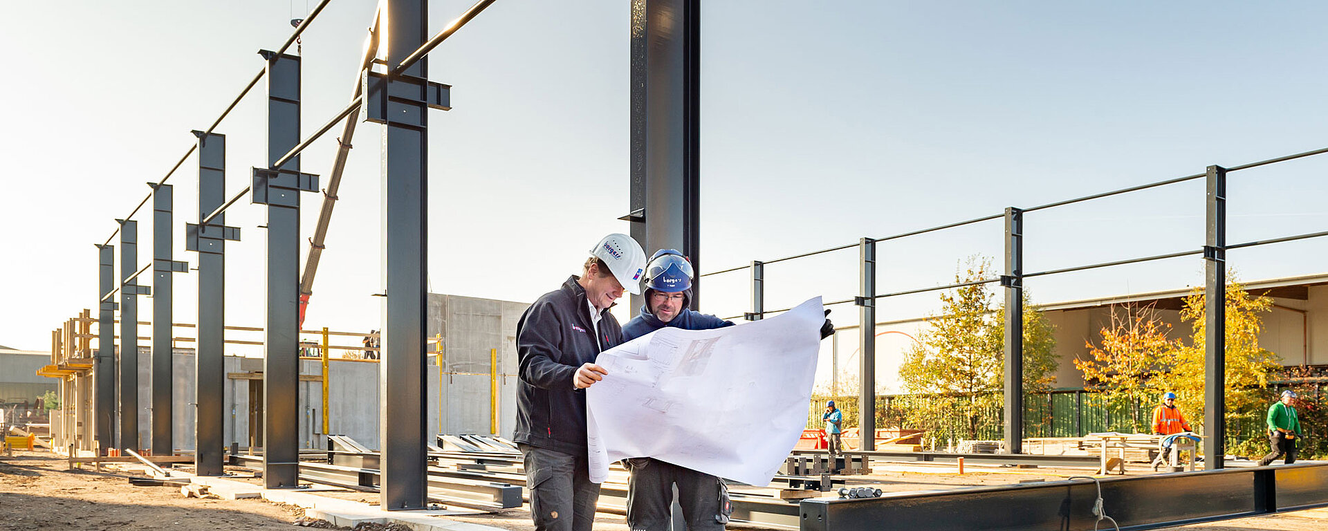 Zwei Bauarbeiter in Schutzkleidung betrachten Baupläne auf einer Baustelle mit Stahlträgern