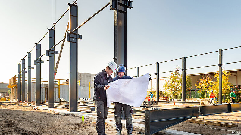 Zwei Bauarbeiter in Schutzkleidung betrachten Baupläne auf einer Baustelle mit Stahlträgern