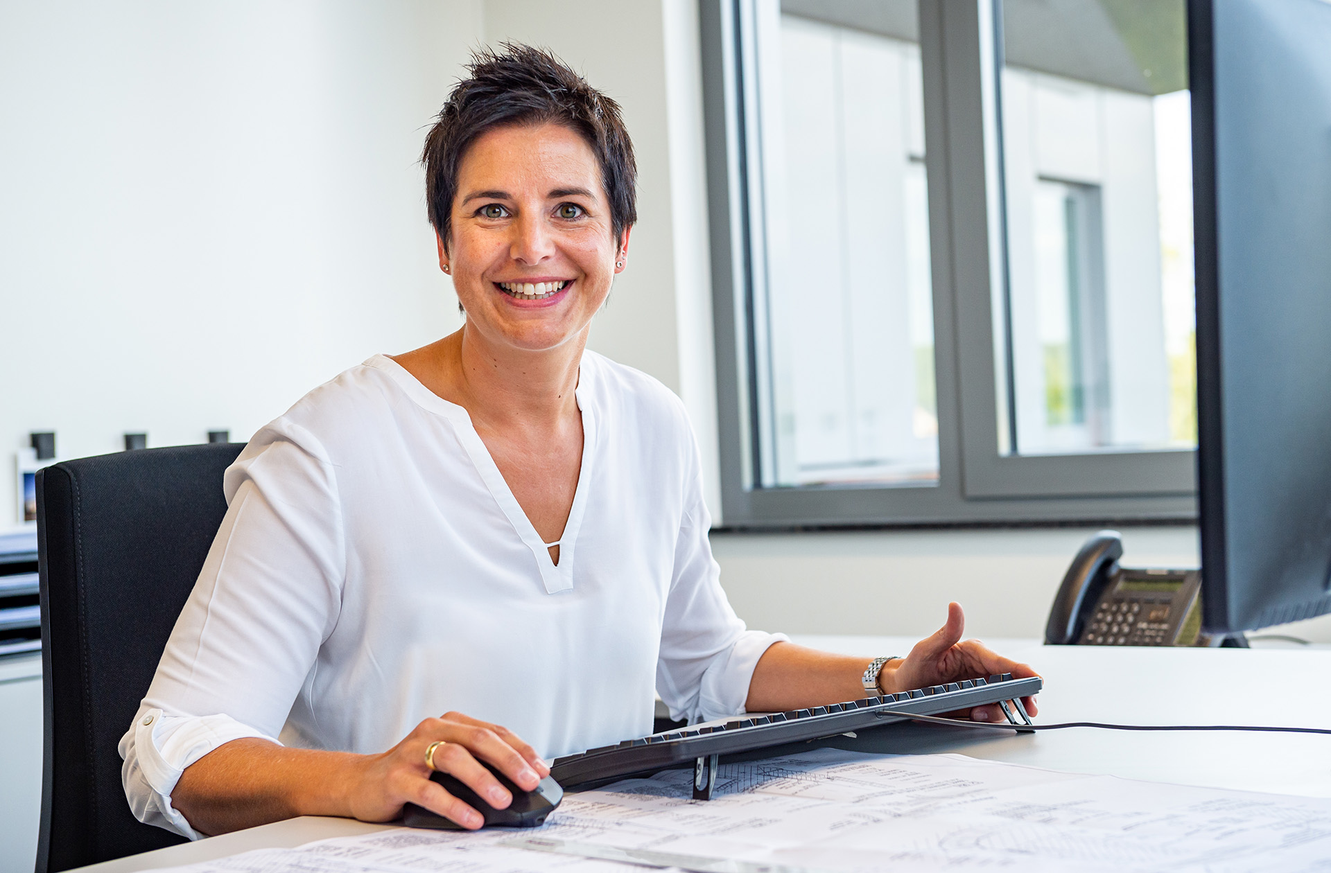 Borgers Mitarbeiterin mit kurzem, dunklem Haar und einem weißen Hemd lächelt in einem Büro mit Bauplänen und einem Computer