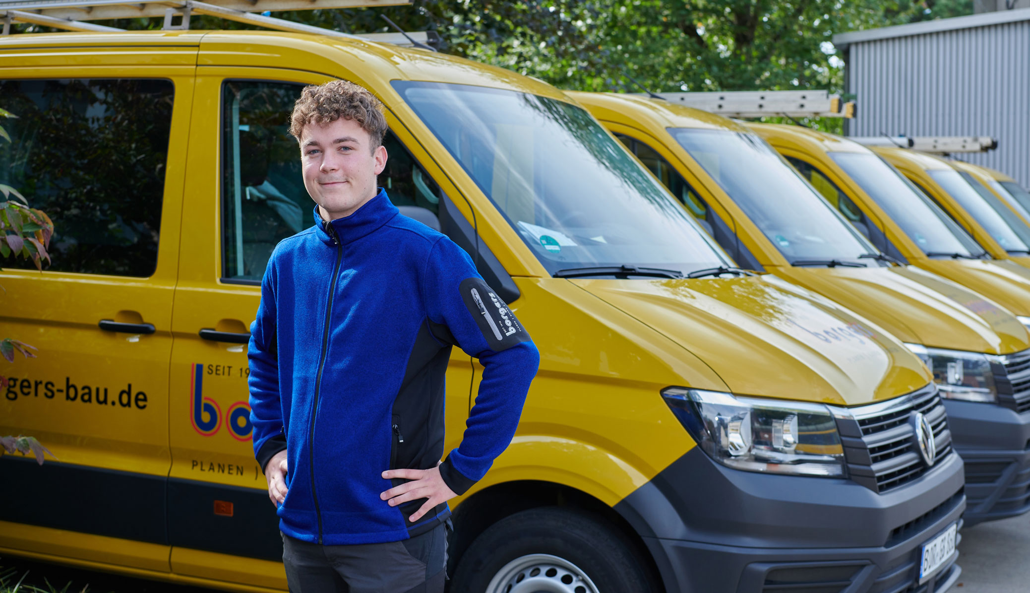 Maurer-Azubi der Borgers GmbH in blauer Jacke, der vor einer Reihe gelber Transporter steht