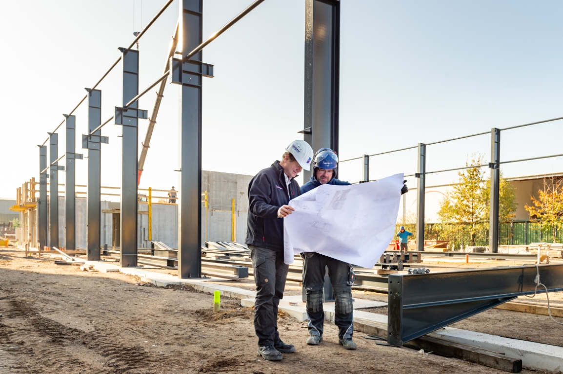 Zwei gewerbliche Mitarbeiter der Borgers GmbH auf der Baustelle. Sie schauen gemeinsam auf einen Plan. Im Hintergrund ist Stahlbau zu sehen.